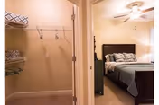 View into a small walk-in closet on the left and a furnished bedroom with a bed, ceiling fan, and nightstand visible on the right.