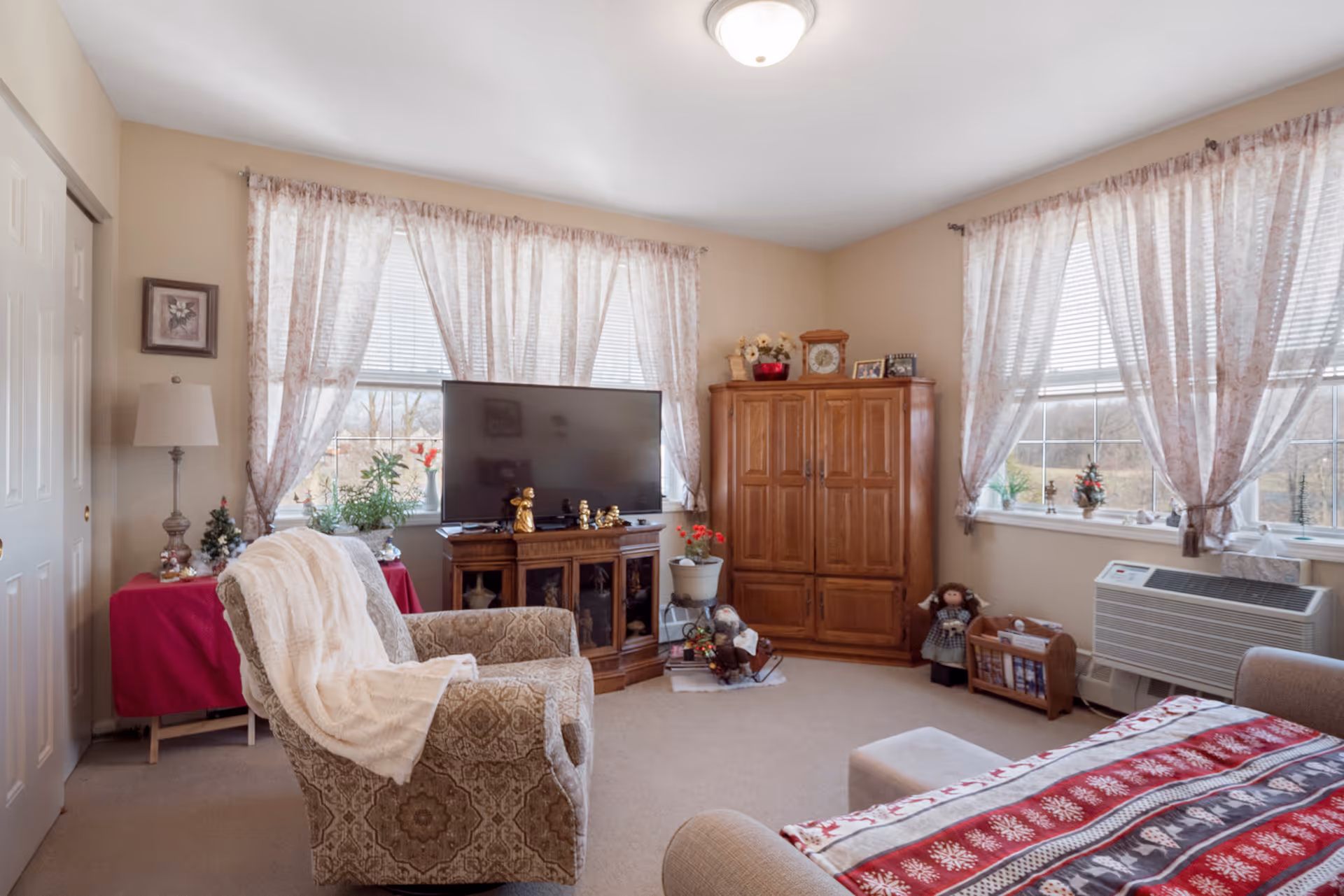 Cozy living room with an upholstered armchair, TV on a wooden stand, a wooden armoire and large windows with sheer curtains.