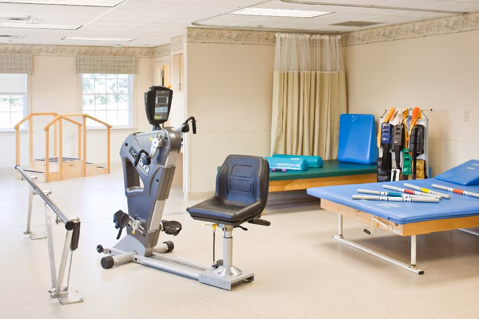 Therapy room with exercise equipment including a recumbent exercise bike, parallel bars, therapy tables, mats and resistance bands.