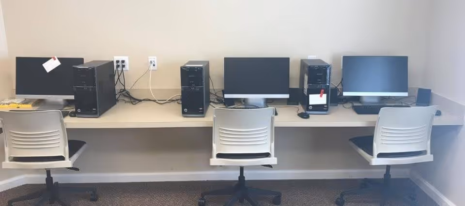 A small computer workstation area with three desktop computers and three office chairs arranged along a wall with beige paint. Each computer has a monitor, keyboard, and tower, with visible cables plugged into wall outlets above the desk.