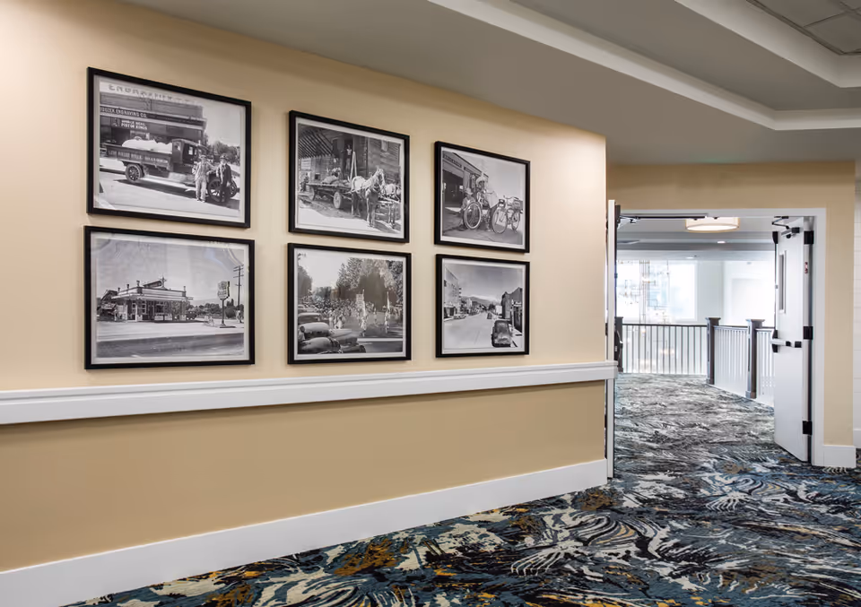 A hallway in Aspen Ridge Residences of Lehi featuring six black and white framed historical photographs on a beige wall. The hallway has patterned carpet in shades of blue, gray, and gold, and an open door leading to a bright area with railing and windows.