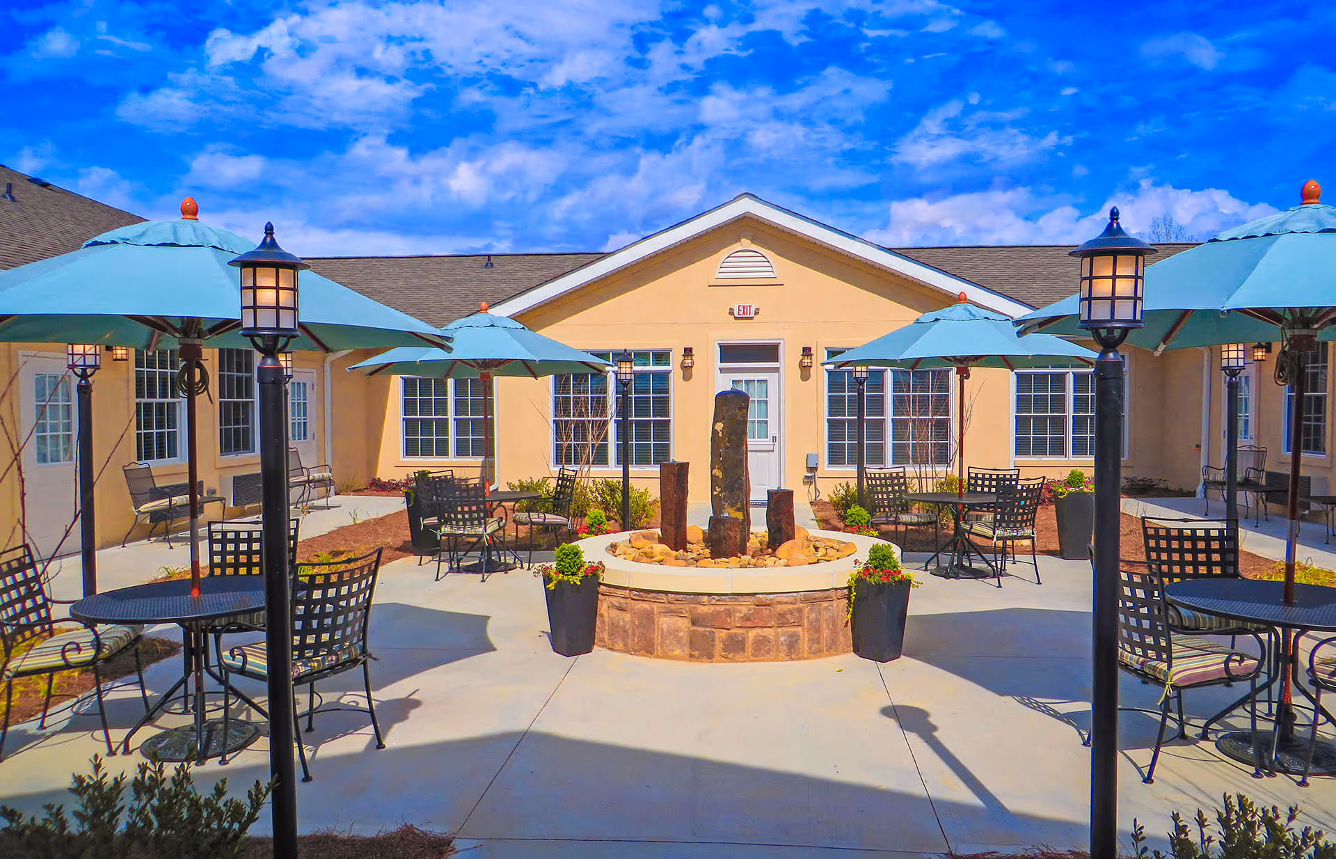 Outdoor courtyard area at Benton House of Grayson featuring several black metal tables and chairs with blue umbrellas, decorative lamp posts, potted plants, and a central stone water fountain, all under a partly cloudy blue sky.