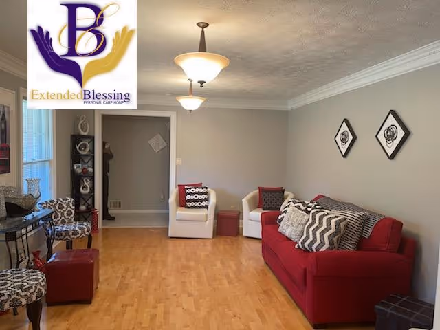 A living room with a red sofa adorned with patterned cushions, two white armchairs with decorative pillows, and two patterned chairs. The room has light wood flooring, beige walls with two framed artworks, and two ceiling light fixtures. A small table with decorative items is placed near the window.