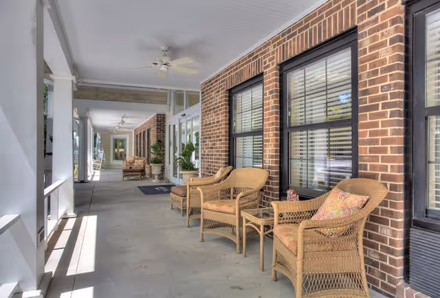 A covered outdoor porch area with wicker chairs and small tables arranged along a brick wall with windows. The porch has ceiling fans and potted plants, creating a comfortable seating space.