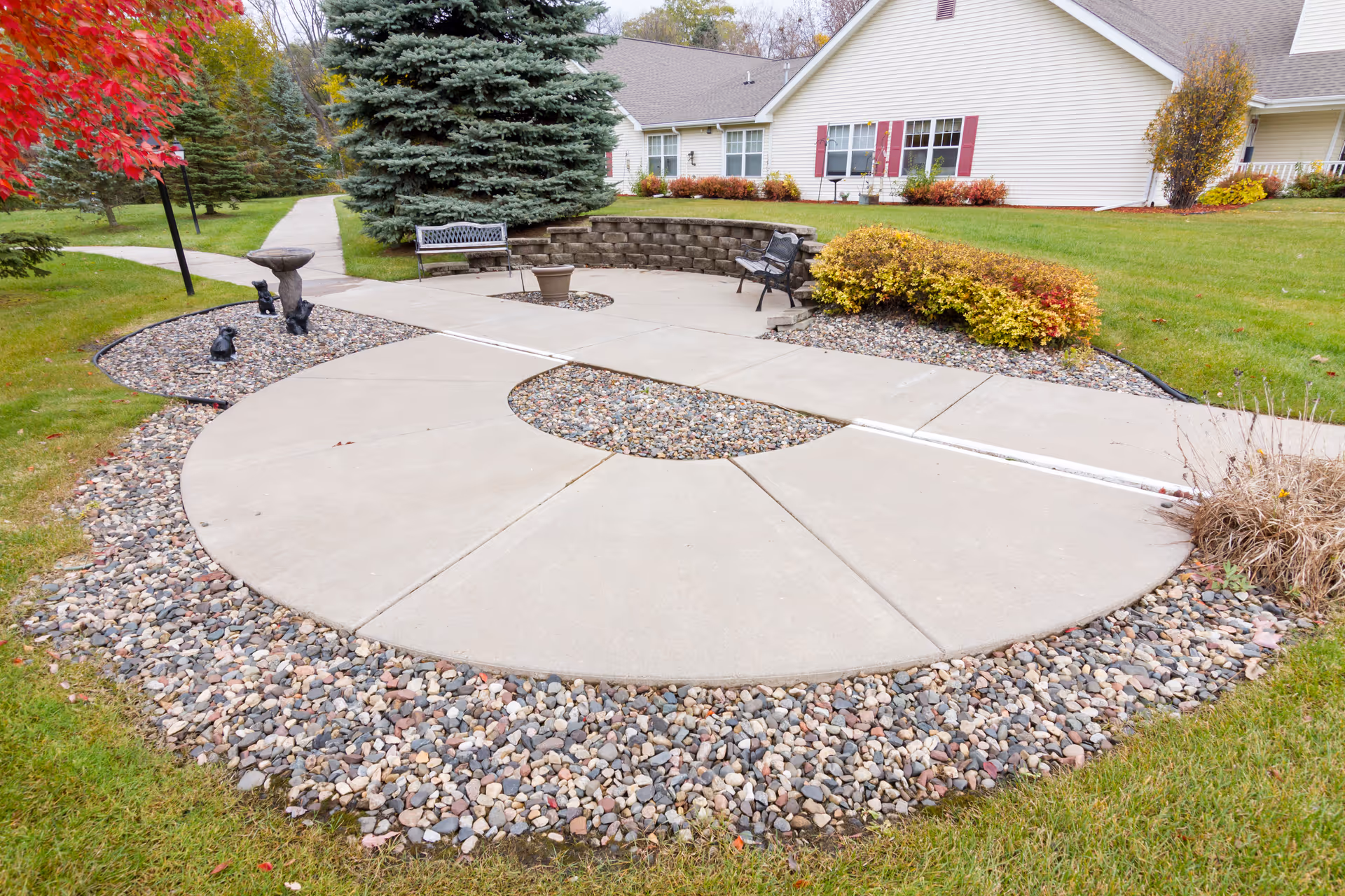 Circular concrete patio and landscaped seating area with benches and shrubs in front of a single-story residential building.