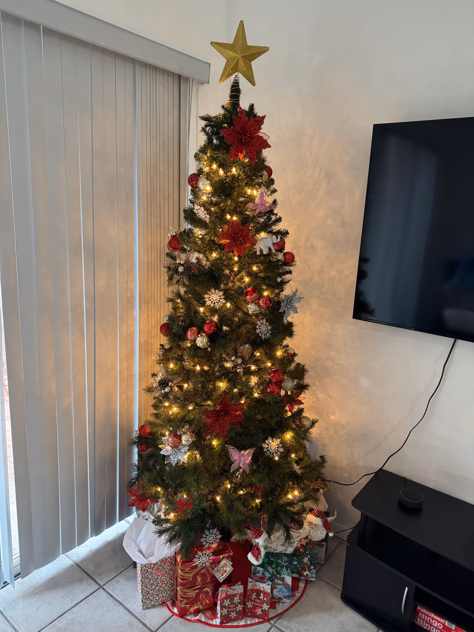 A decorated Christmas tree with lights, red and gold ornaments, snowflakes, and a gold star on top, standing next to vertical blinds and a wall-mounted flat screen TV. Several wrapped presents are placed under the tree on a tiled floor.