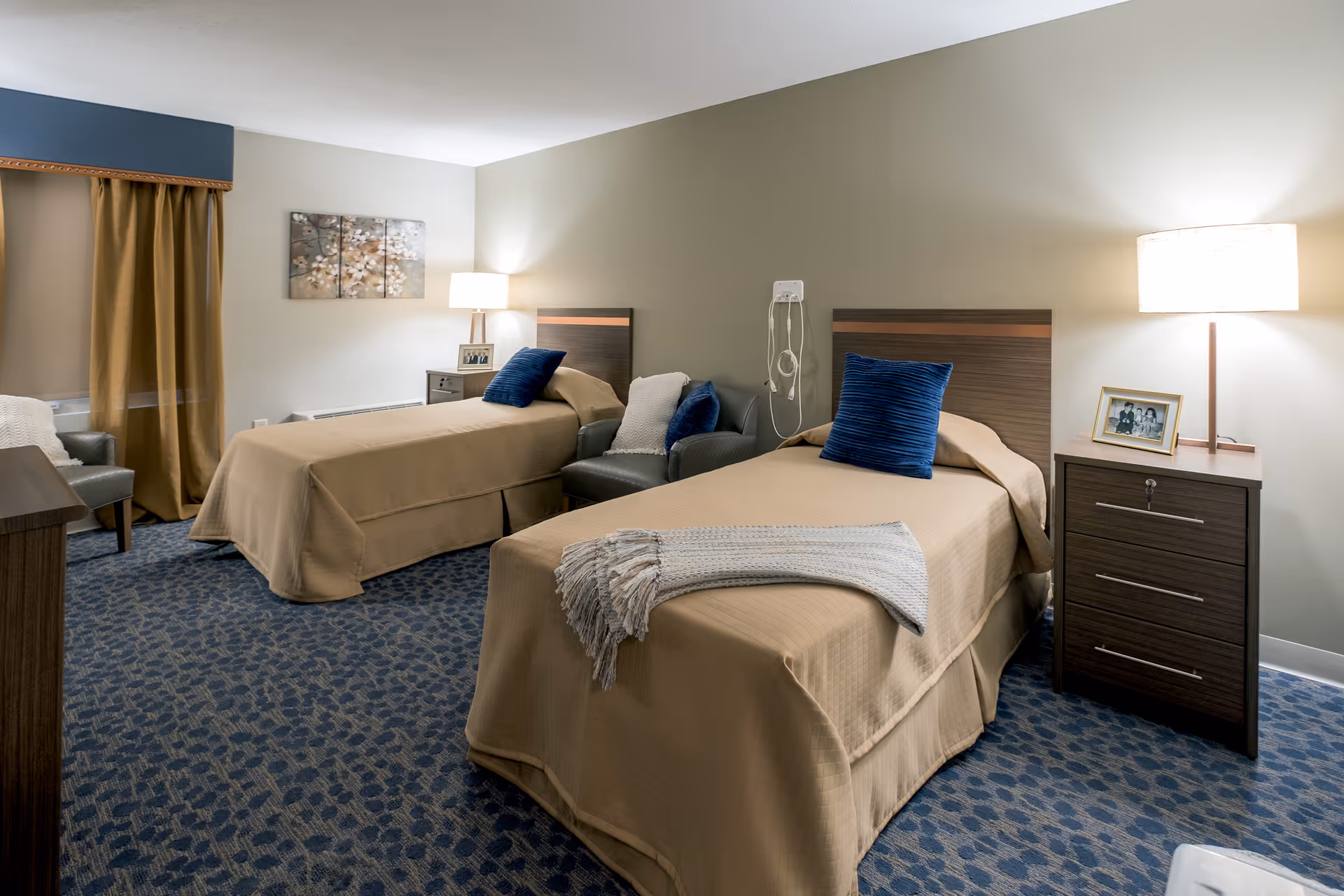 A senior living facility bedroom with two single beds covered in beige bedspreads, each with a blue pillow. Between the beds is a small nightstand with a lamp and a framed photo. There are two armchairs with blue pillows next to the beds. The room has patterned blue carpet, beige walls, a window with brown curtains, and a piece of floral artwork on the wall.