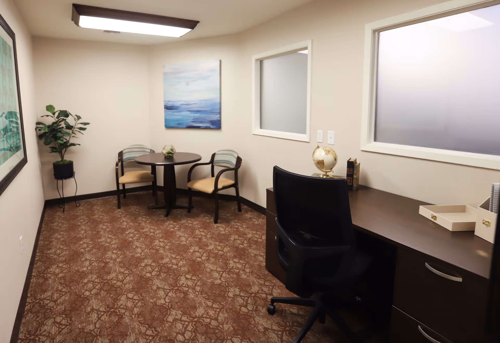 A small office or meeting room with a brown patterned carpet, beige walls, and a ceiling light. There is a dark wooden desk with a black office chair, a globe, and some books on the desk. Against the far wall, there is a round table with two chairs and a small plant on the table. A potted plant stands in the corner, and two framed artworks hang on the walls. Two frosted windows are set into the wall above the desk.