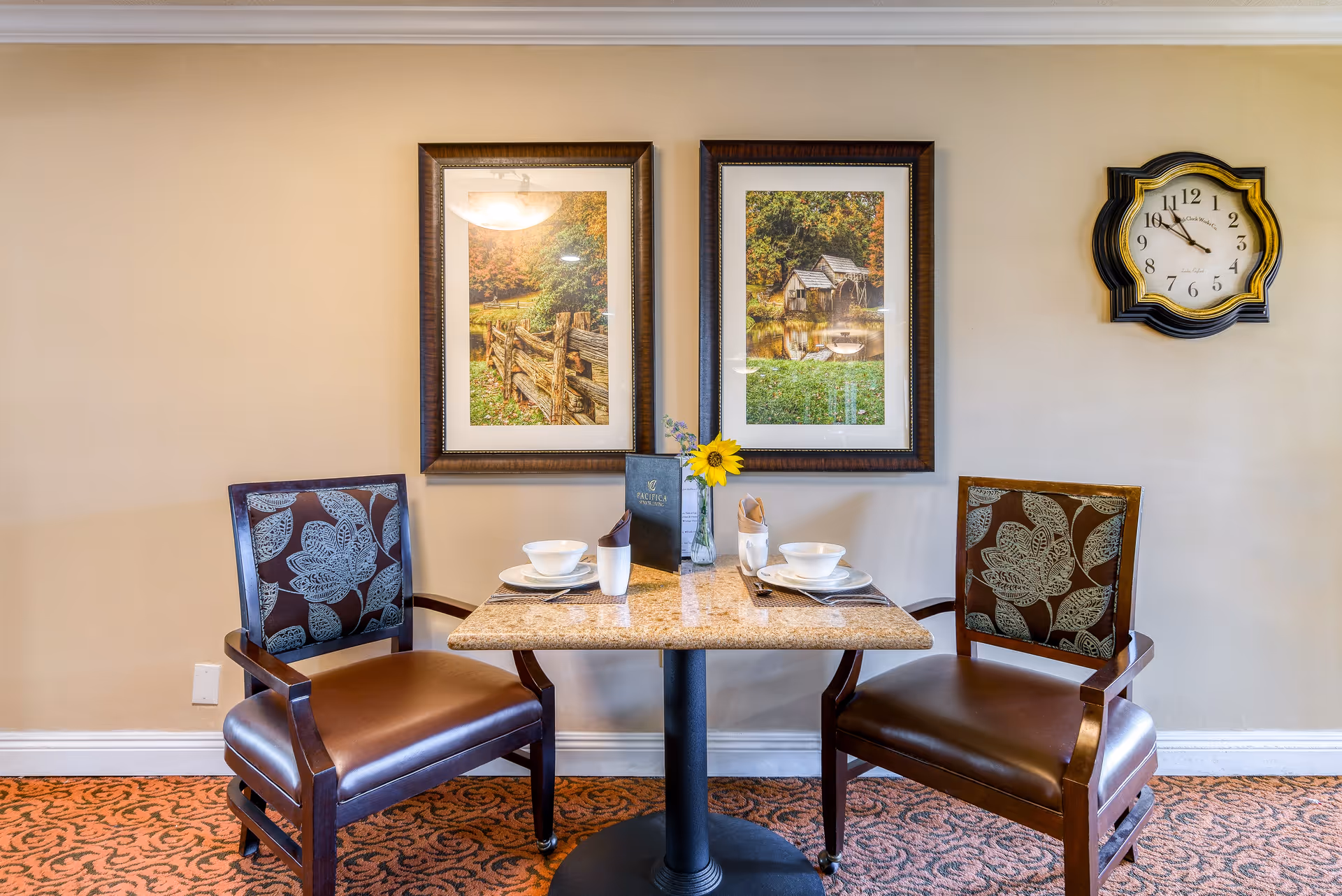 A small dining table set for two with two chairs featuring floral upholstery. The table has a granite top and is set with white bowls, plates, cups, and silverware. A small vase with a sunflower and a menu are placed in the center. Two framed landscape pictures hang on the beige wall behind the table, and a decorative wall clock is mounted to the right.