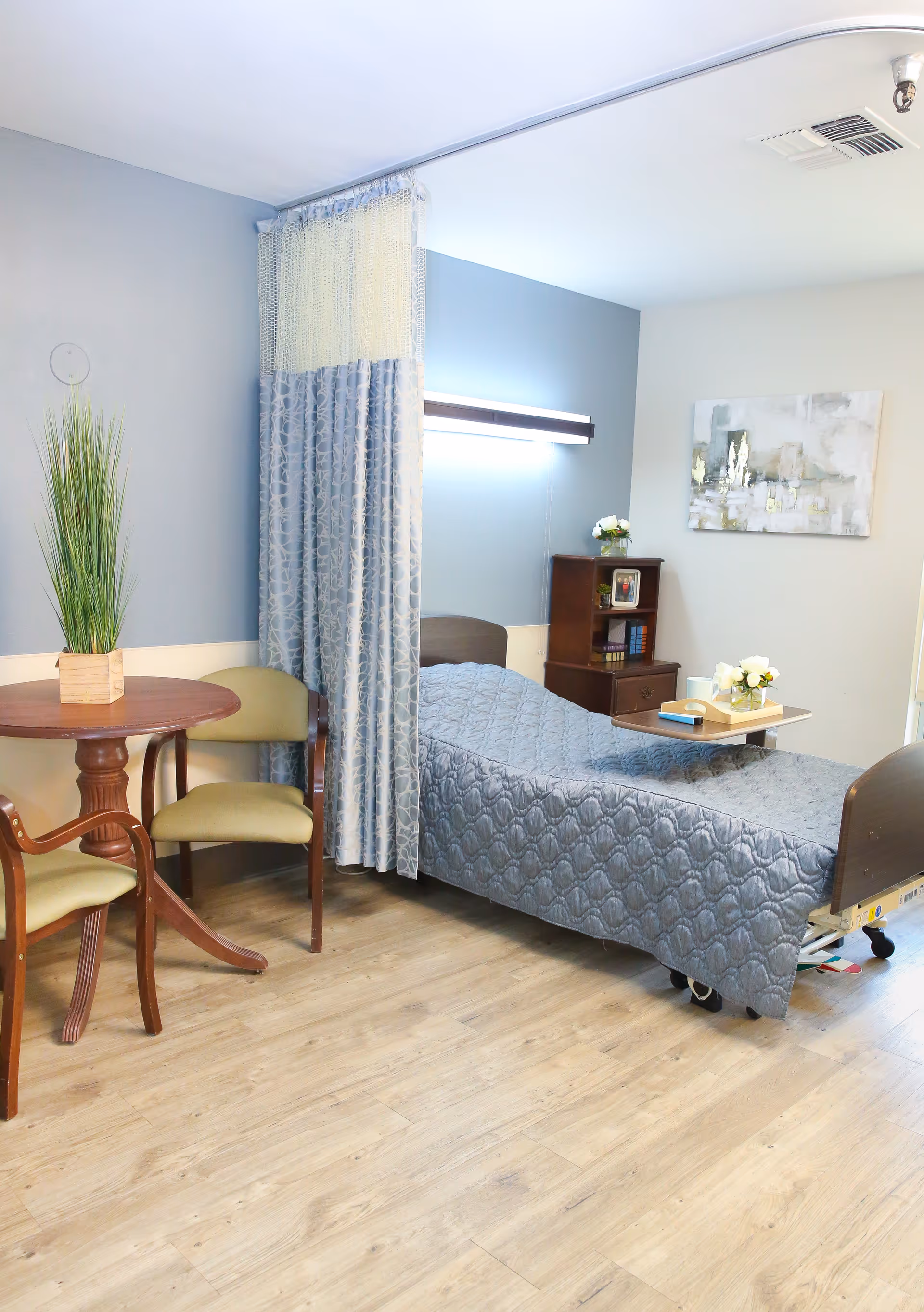 A tidy senior facility patient room with a single bed, bedside table and tray, privacy curtain, small round table with chairs, and wall decor.