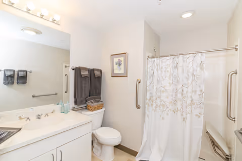 A clean and well-lit bathroom featuring a white vanity with a sink, a large mirror with lights above it, a toilet with folded dark gray towels on a rack above it, a framed floral picture on the wall, and a shower area with a white curtain decorated with a subtle leaf pattern and a foldable shower seat inside.