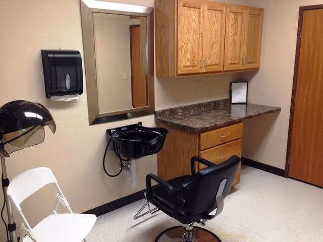 Interior view of a small salon or grooming area in an assisted living facility, featuring a black salon chair in front of a black wash basin attached to the wall, a large mirror above the basin, wooden cabinets with a countertop, a white folding chair, and a black hair dryer hood.