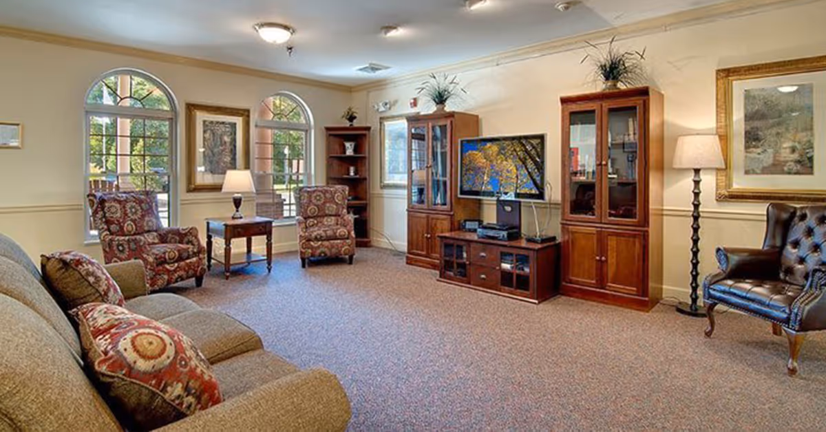 A cozy senior living facility common area with a beige sofa adorned with patterned cushions, two patterned armchairs, a wooden side table with a lamp, a wooden entertainment center with a TV displaying a nature scene, and a leather armchair. The room has arched windows letting in natural light, framed artwork on the walls, and carpeted flooring.