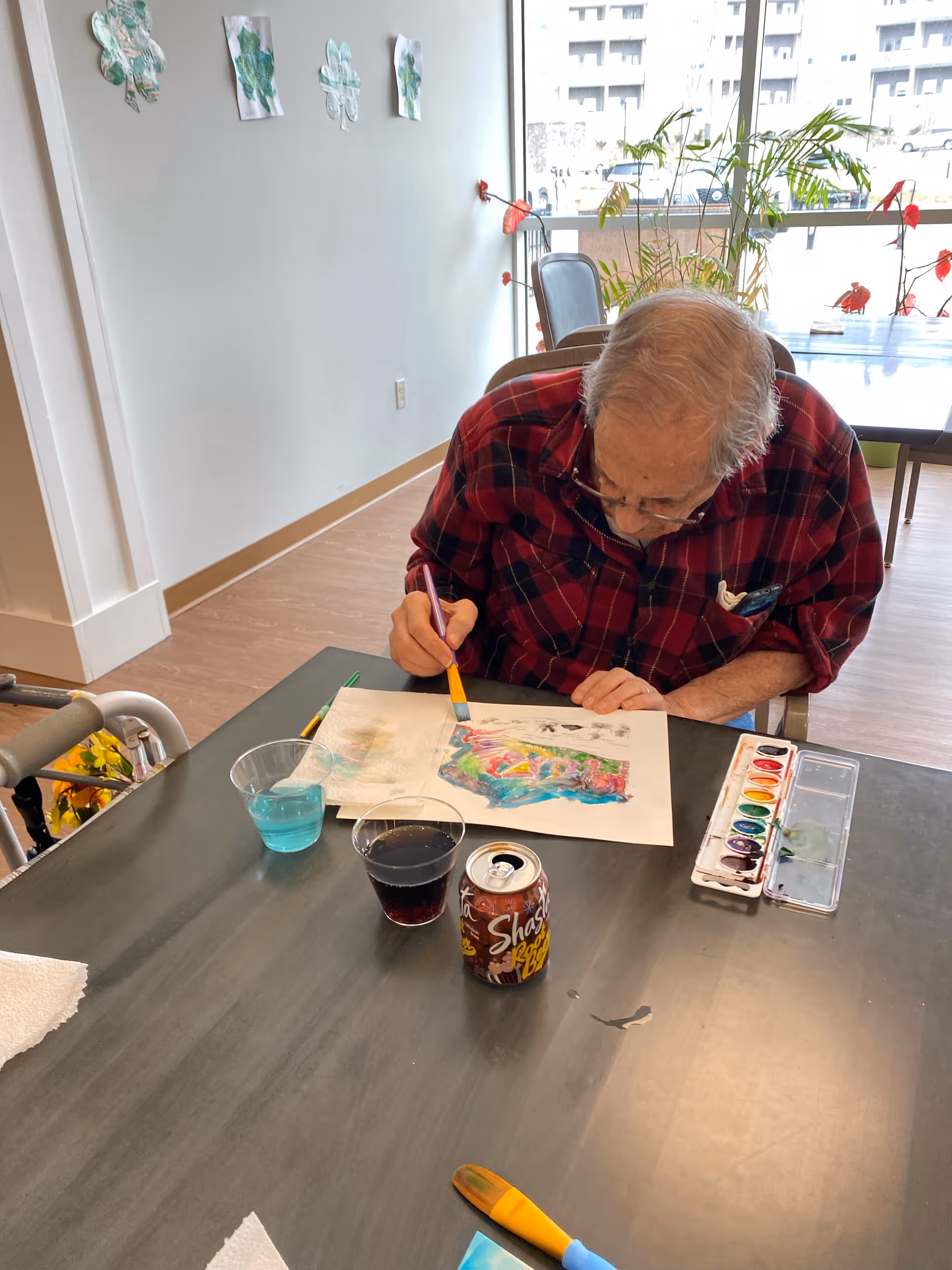 An elderly man wearing a red plaid shirt is sitting at a table painting with watercolors. On the table are a watercolor palette, a paintbrush, a can of Shasta root beer, a cup with blue liquid, and a cup with a dark beverage. The room has light-colored walls with some leaf-shaped decorations and a large window with plants and a view of a building outside.