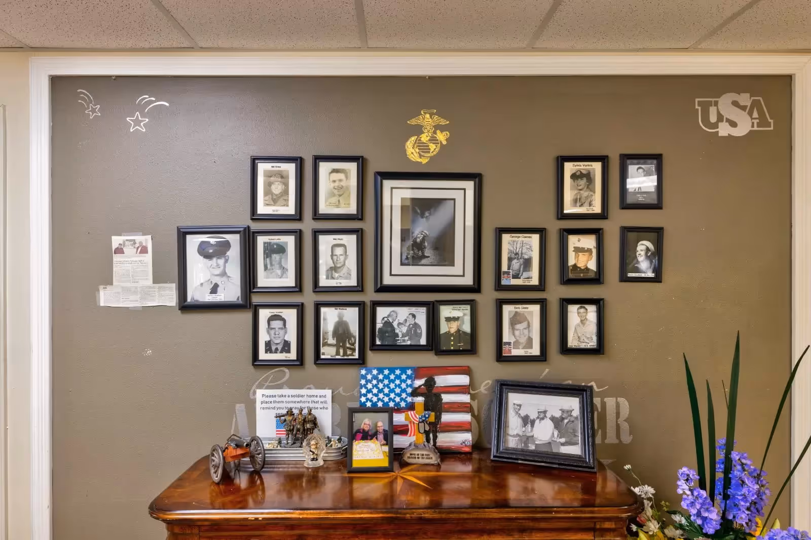 A wall display featuring framed black and white photographs of military personnel, a golden Marine Corps emblem, and a USA decal. Below the photos is a wooden table with patriotic decorations including a small American flag, a toy cannon, figurines of soldiers, and framed pictures. There are also some purple flowers on the right side of the table.