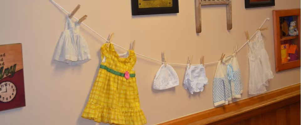 A wall display featuring a clothesline with small baby clothes and accessories hanging on clothespins, including a yellow dress, white hats, and other small garments. The wall has wooden paneling at the bottom and framed decorations.