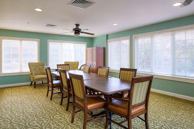 A bright room with large windows covered by white blinds, featuring a long wooden table surrounded by eight wooden chairs with striped upholstery. Two upholstered armchairs are placed near the windows, and a ceiling fan is mounted on the light green ceiling. The floor is covered with a patterned carpet in shades of green and beige.