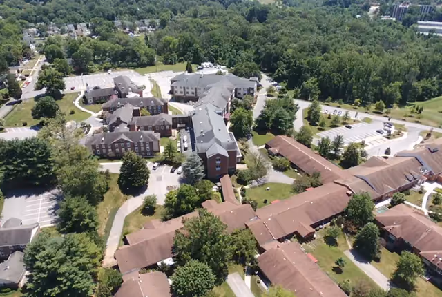 Aerial view of the Resorts of Augsburg senior living facility showing multiple connected buildings with brown and gray roofs surrounded by trees, parking lots, and green spaces.
