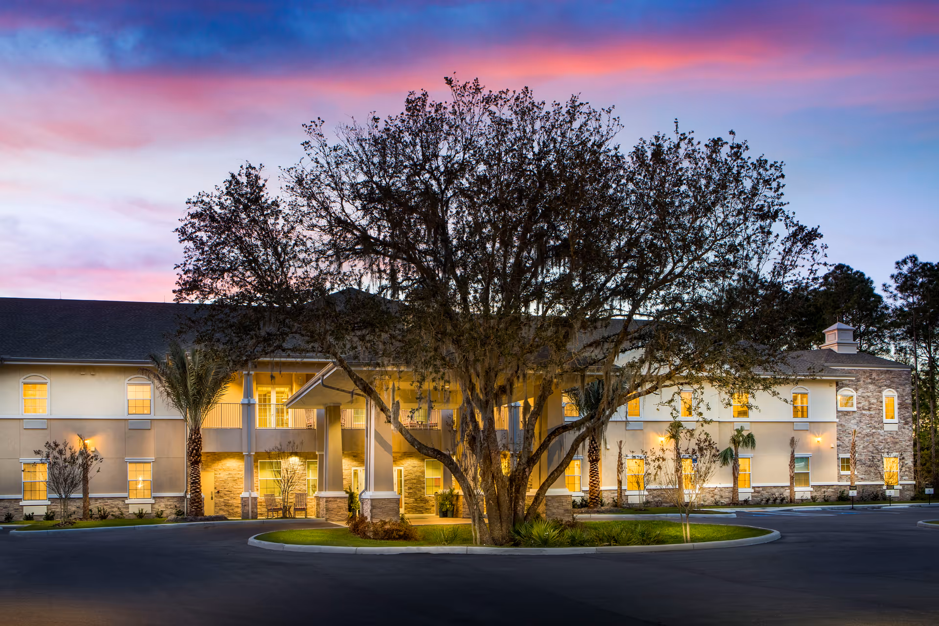 Front exterior of a two-story senior living building lit at dusk with a large tree in the circular driveway.