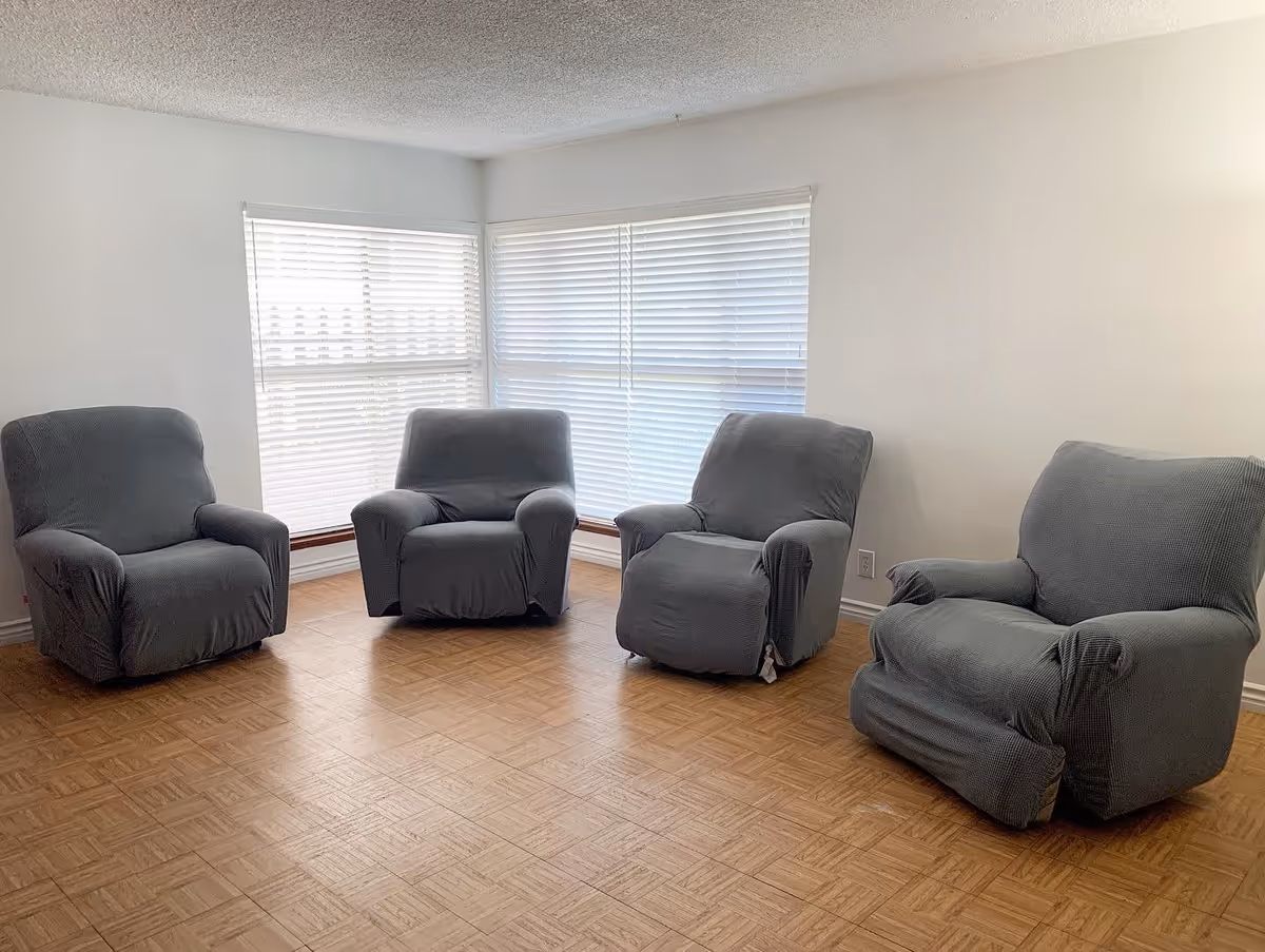 A room with four gray upholstered armchairs arranged in a semi-circle on a wooden parquet floor. The room has white walls and two large windows with white blinds letting in natural light.