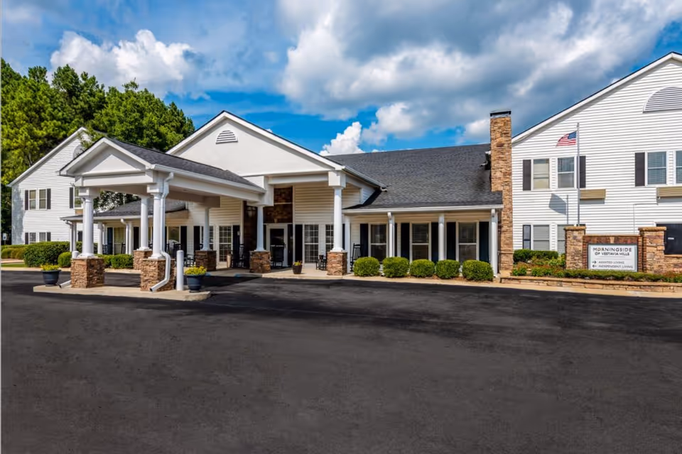 Exterior front view of Morningside of Vestavia Hills senior living facility showing a white building with a covered entrance supported by stone pillars, neatly trimmed bushes, an American flag, and a sign indicating assisted living and independent living services under a partly cloudy blue sky.