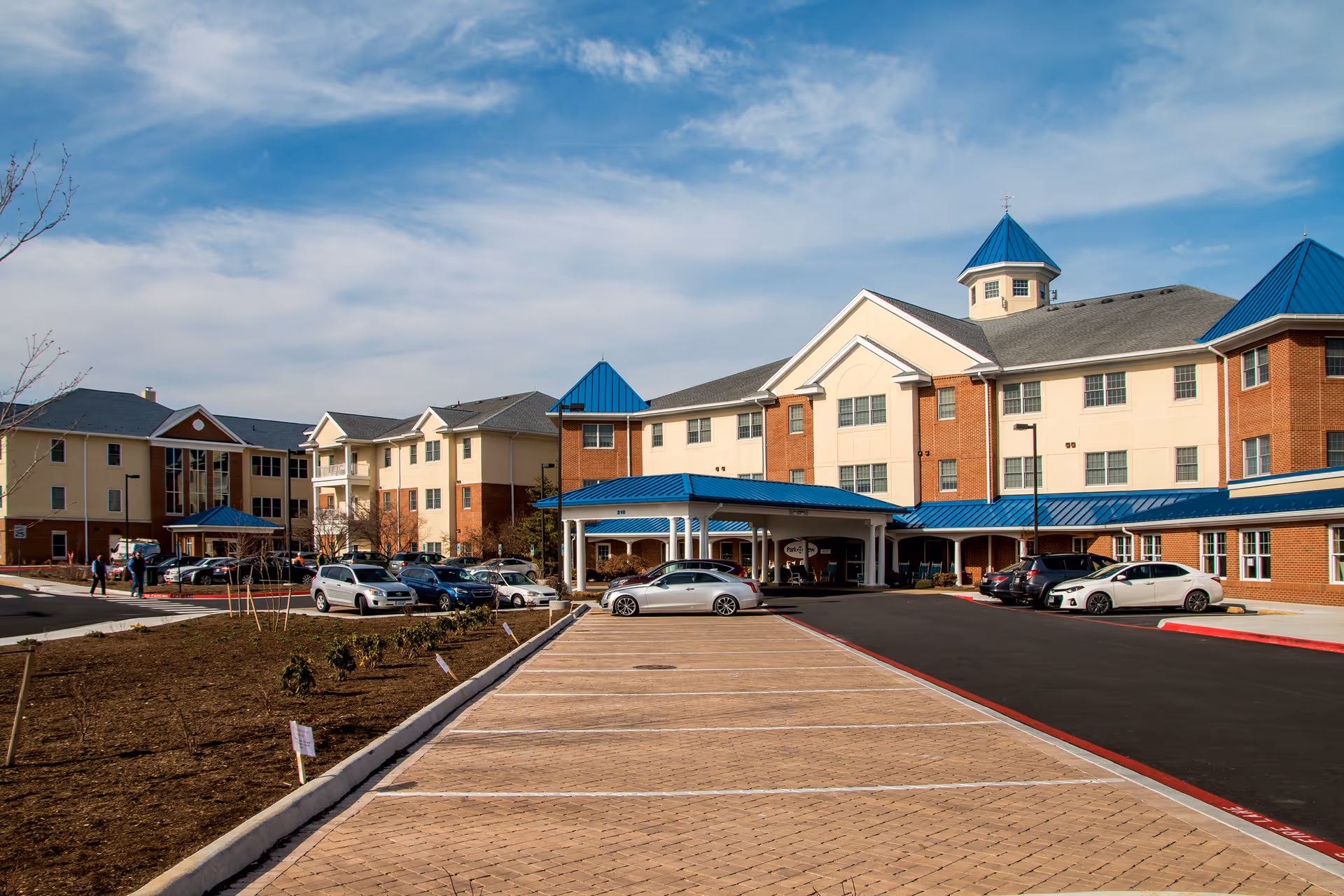Exterior view of Bridgewater Retirement Community showing a large multi-story building with beige and brick walls and blue roofs. There is a covered entrance with cars parked in front and a paved parking lot extending towards the foreground. The sky is partly cloudy.