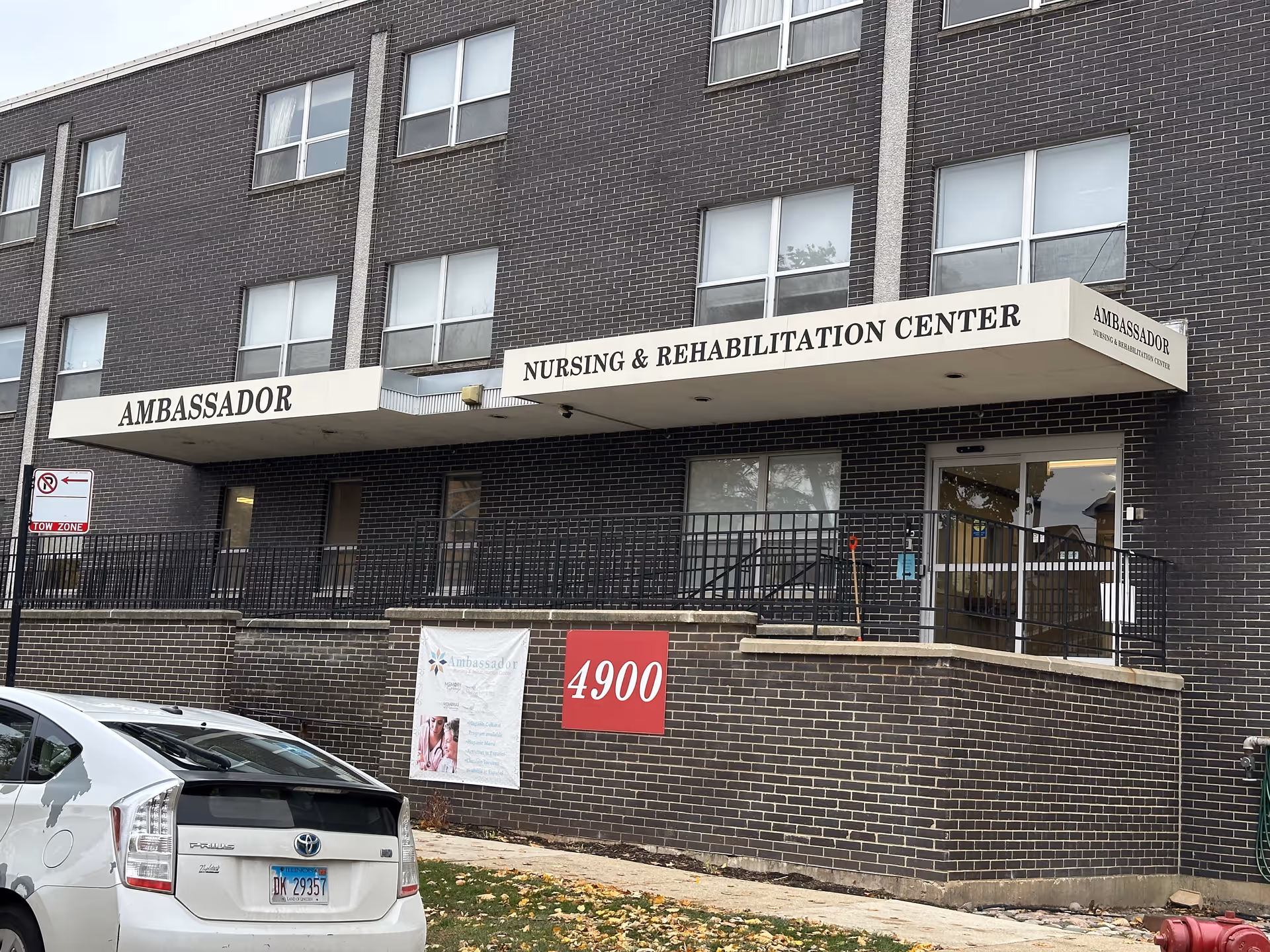 Exterior view of the Ambassador Nursing & Rehabilitation Center building with dark brick walls, multiple windows, and a white awning displaying the facility's name. A white Toyota Prius is parked in front, and a red sign with the number 4900 is mounted on the brick wall near the entrance.