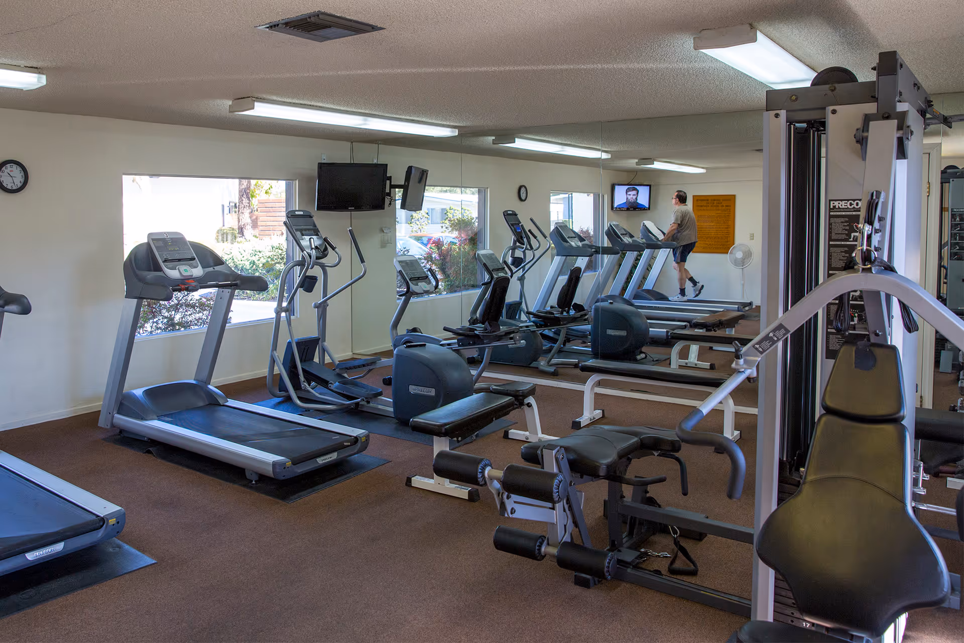 Interior view of a senior living community fitness room with several treadmills, elliptical machines, and weight training equipment. A man is walking on one of the treadmills, and there are windows and mirrors along the walls.