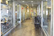 A long, well-lit hallway with glass walls on both sides, showing rooms with medical equipment and chairs lined up along the right side of the corridor. The floor has a polished, reflective surface and the ceiling has recessed lighting.