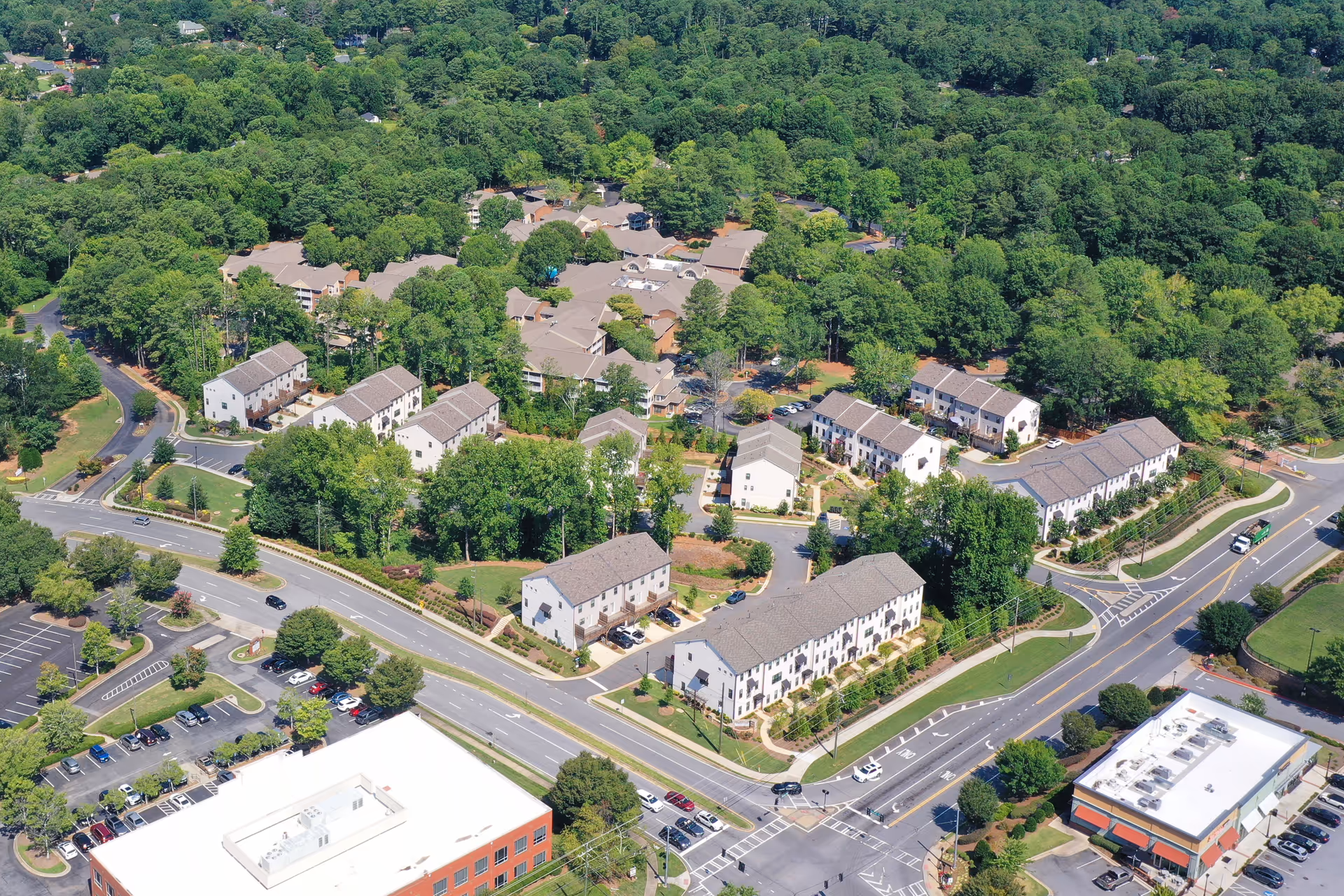 Aerial view of Brookdale Chambrel Roswell senior living facility surrounded by dense green trees. The complex consists of multiple residential buildings with beige and white exteriors and gray roofs, arranged along curved roads. Adjacent to the facility are parking lots and commercial buildings, with roads and intersections visible in the foreground.