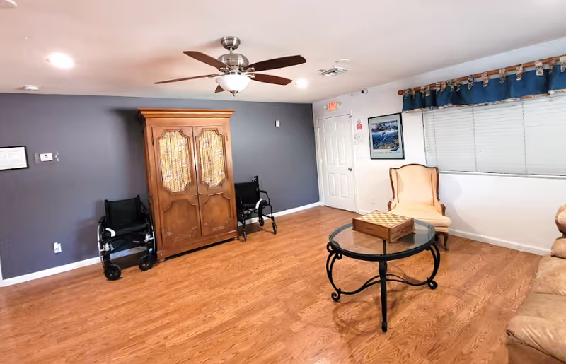 Bright common living room with hardwood floors, a ceiling fan, an armoire, seating including wheelchairs and an armchair, and a glass-top coffee table.