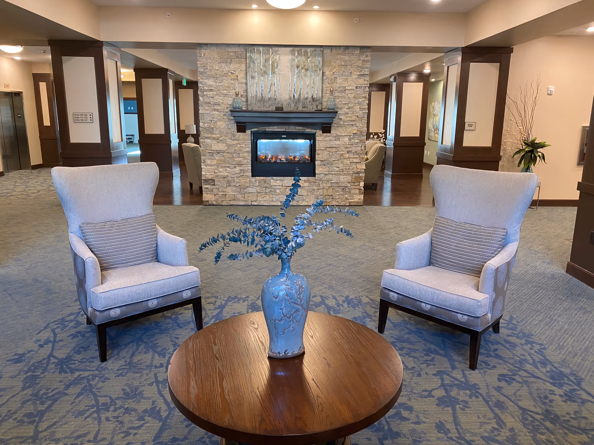 A cozy seating area with two light gray armchairs facing each other across a round wooden table with a blue vase holding decorative branches. Behind the seating area is a stone fireplace with a modern electric fire, set in a spacious room with carpeted floor and wooden pillars.