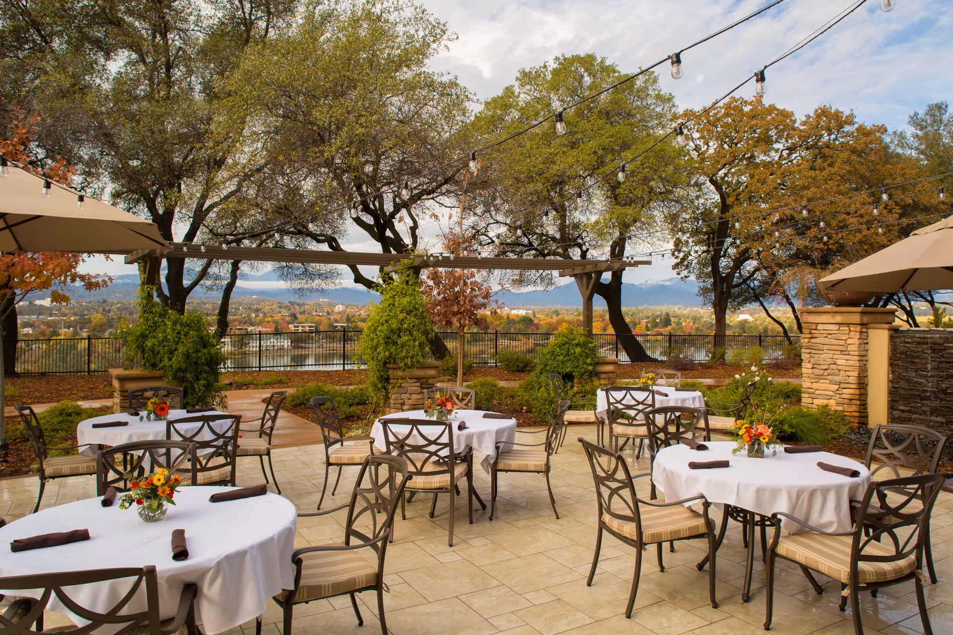 Outdoor patio area with round tables covered in white tablecloths, each set with a small floral centerpiece and brown napkins. The patio has metal chairs with beige cushions and is surrounded by trees and greenery. String lights are hung overhead, and there is a scenic view of a lake and distant mountains under a partly cloudy sky.
