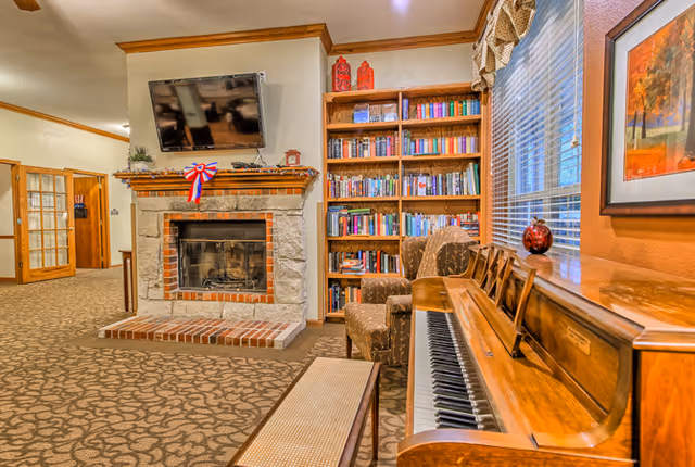 A cozy living area in Trail Creek Place Assisted Living featuring a stone fireplace with a mounted flat-screen TV above it, a wooden bookshelf filled with books, two patterned armchairs, a wooden piano with a bench, and a window with blinds and a valance. The room has carpeted flooring and warm-toned walls with crown molding.
