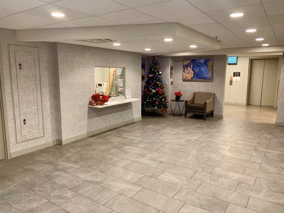 Interior view of a senior living facility lobby area with gray tiled floor and light gray walls. There is a Christmas tree decorated with colorful lights and ornaments near a small table with a red poinsettia plant. A single armchair is placed next to the table. On the wall above the chair is a framed abstract artwork. To the right, there is an open hallway leading to two elevator doors. On the left wall, there is a reception window with a counter and holiday decorations. The ceiling has recessed lighting.