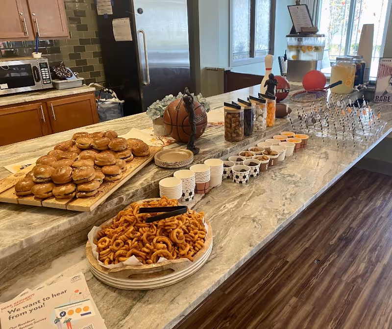 Buffet setup on a marble countertop with sliders, a bowl of curly fries, small cups, and drink dispensers in a communal kitchen area.
