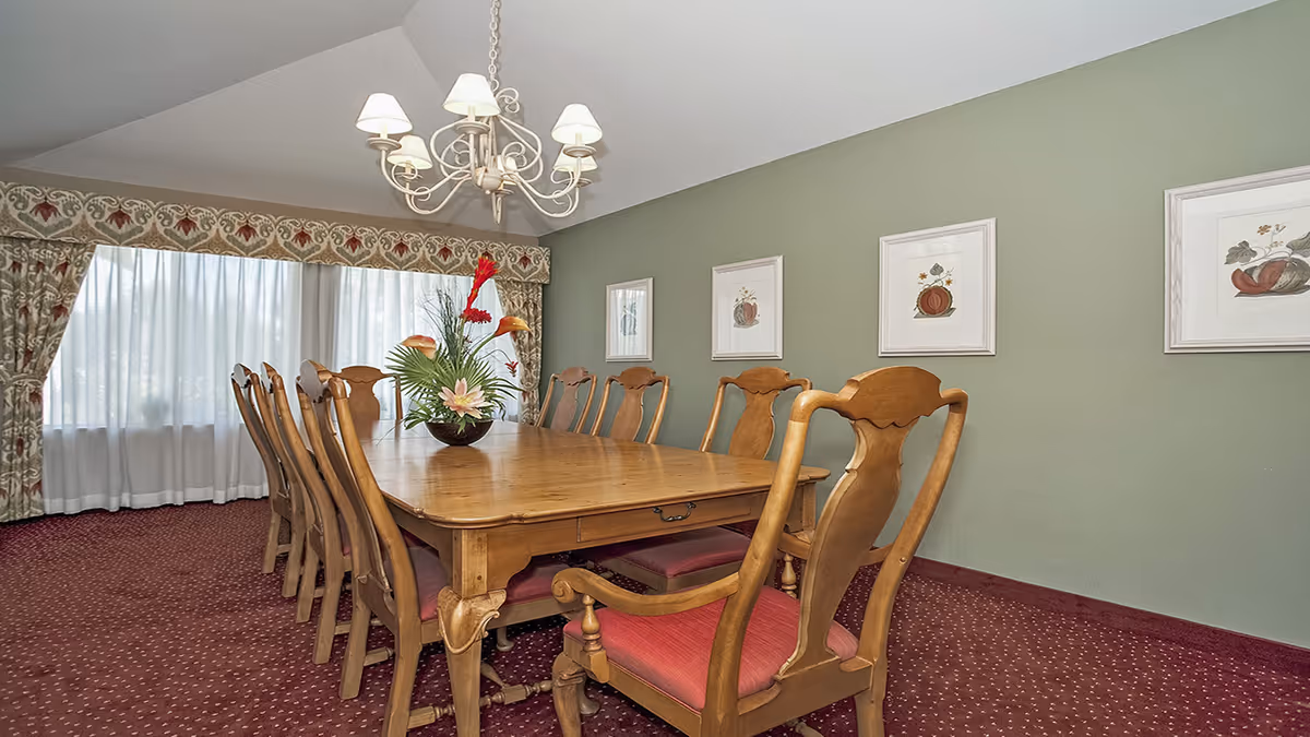 A dining room with a long wooden table surrounded by ten wooden chairs with red cushions. A floral centerpiece is placed on the table. The room has green walls adorned with framed artwork and a large window with patterned curtains letting in natural light. A chandelier with six lamps hangs above the table.