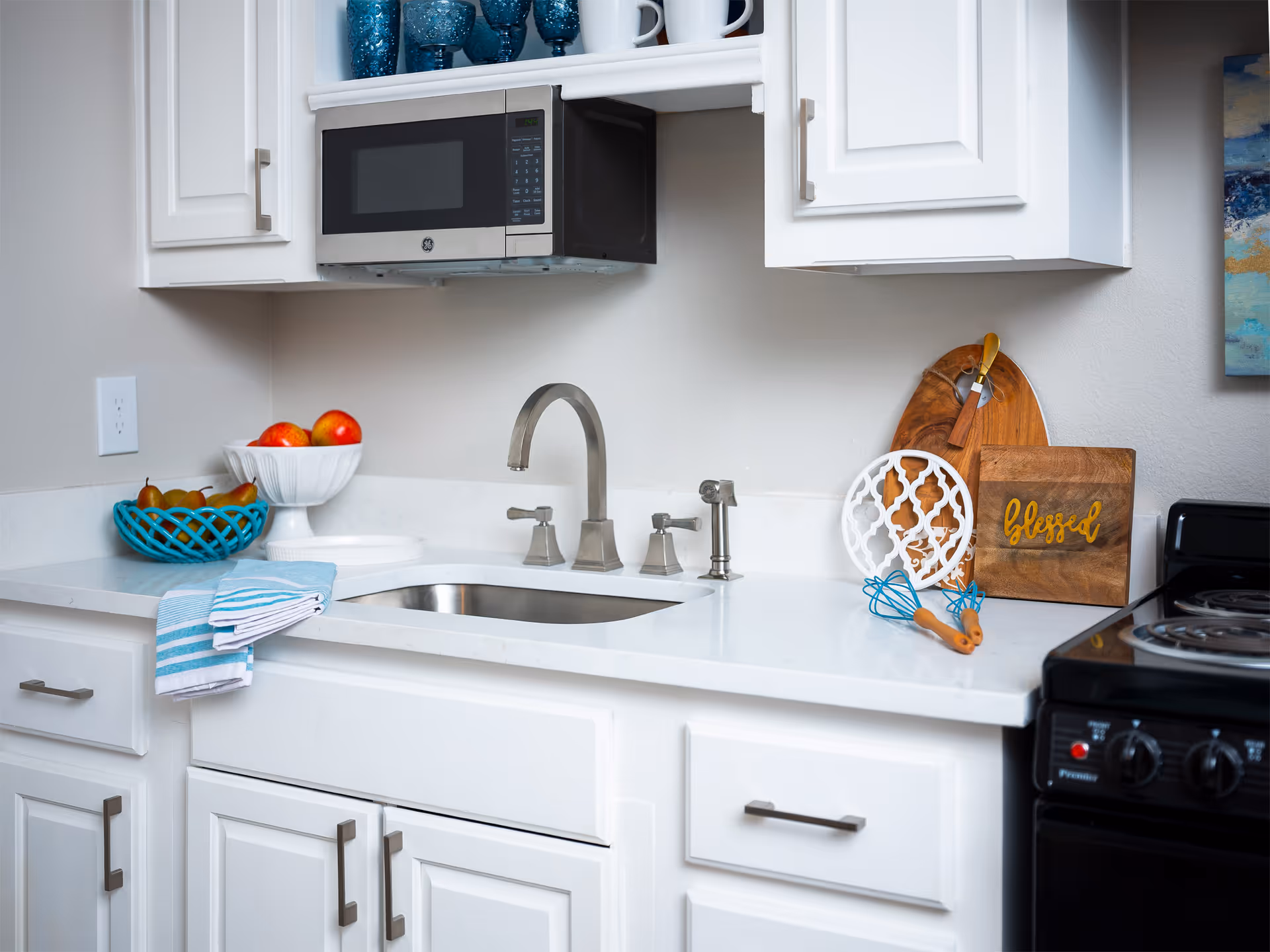 Bright white kitchen countertop with a sink and faucet, overhead microwave, stove, and decorative bowls, cutting boards, and utensils.