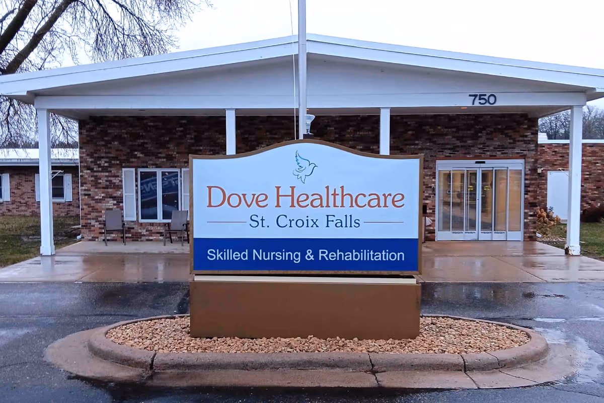 Front exterior view of Dove Healthcare - St. Croix Falls building with a large sign in front that reads 'Dove Healthcare St. Croix Falls Skilled Nursing & Rehabilitation'. The building has a brick facade, white pillars, and automatic glass doors under a covered entrance.