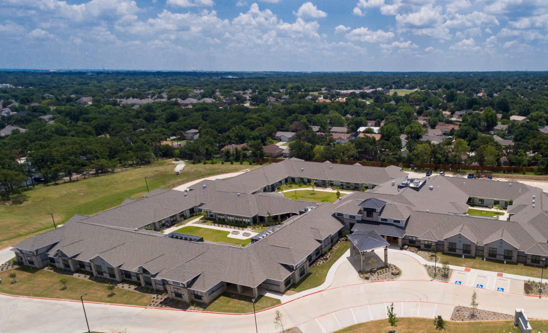 Aerial view of a sprawling assisted living and memory care complex with central courtyards, parking area, and surrounding neighborhood.