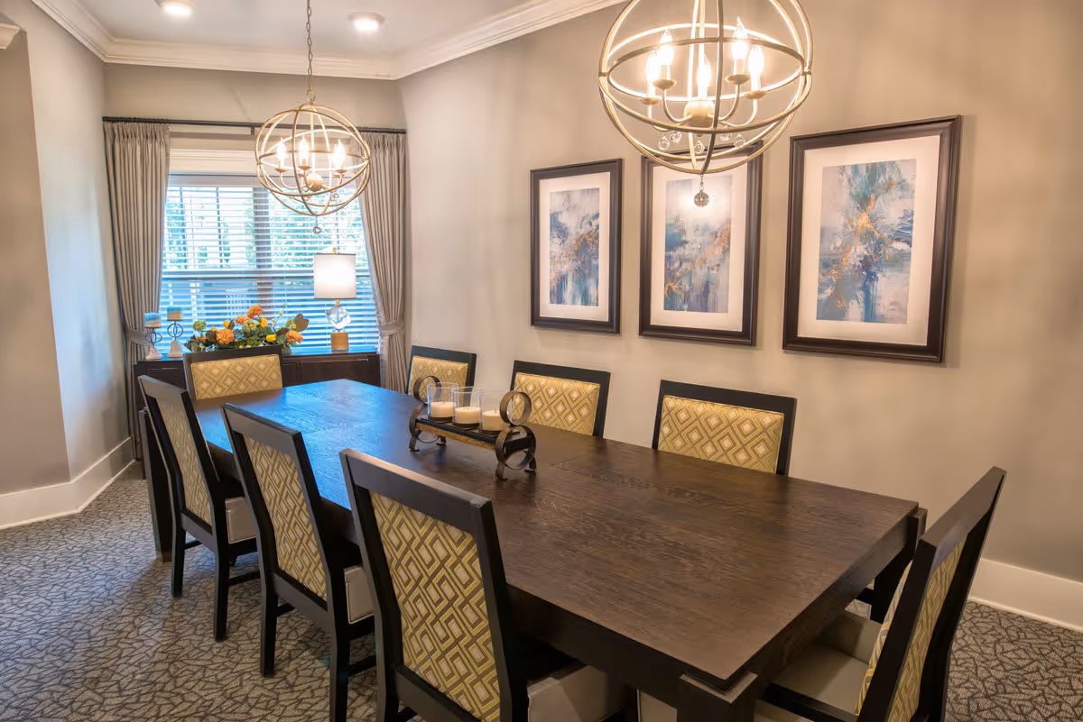 Sunlit dining room with a long dark wood table surrounded by upholstered chairs, two spherical chandeliers, framed artwork on the wall, and a window with curtains.