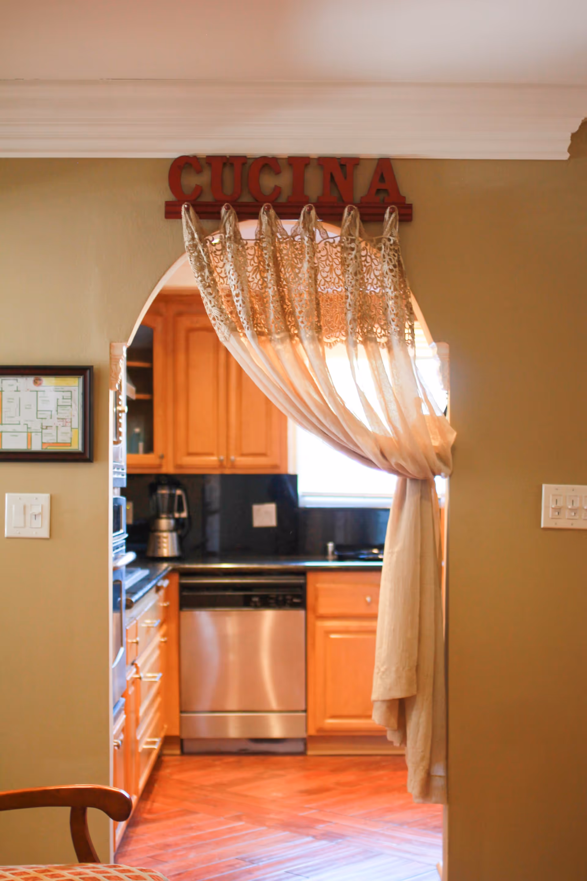 View through an arched doorway with a sheer curtain tied to one side, leading into a kitchen with wooden cabinets, a stainless steel dishwasher, a coffee maker, and a window letting in natural light. Above the doorway is a red sign that reads 'CUCINA'.