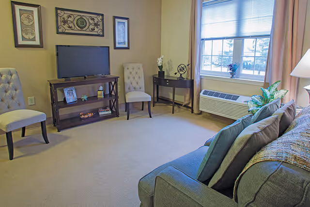 A cozy living room with a gray couch adorned with pillows and a throw blanket, two beige tufted chairs, a small wooden TV stand with a flat-screen TV, decorative items, and framed artwork on the wall. There is a window with beige curtains letting in natural light, a small black side table with a plant and decorative globe, and a floor lamp beside the couch.