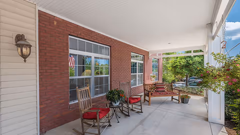 Covered outdoor patio area with two wooden rocking chairs with red cushions, a small round table with a plant, a wooden bench with red and white striped cushions, hanging flower baskets, and a view of trees and parked cars in the background.