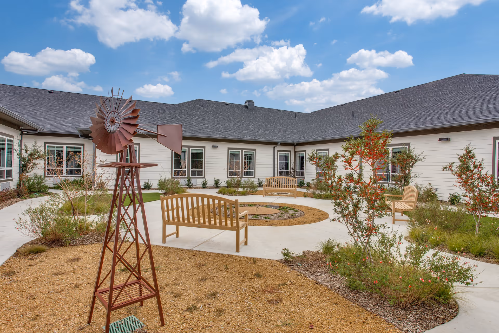 Outdoor courtyard area at Harvest of Aledo Senior Living featuring a small decorative windmill, wooden benches, landscaped plants, and a concrete walkway under a partly cloudy blue sky.