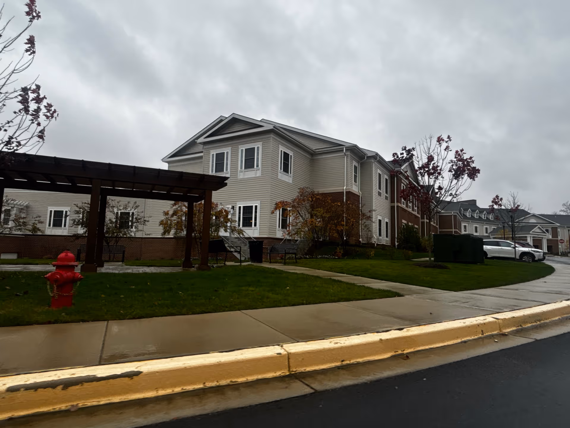 Exterior view of a senior living facility building on a cloudy day. The building has beige siding with white trim and brick accents. There is a wooden pergola structure on the lawn, a red fire hydrant, some small trees with autumn leaves, a sidewalk, and a driveway with parked cars.