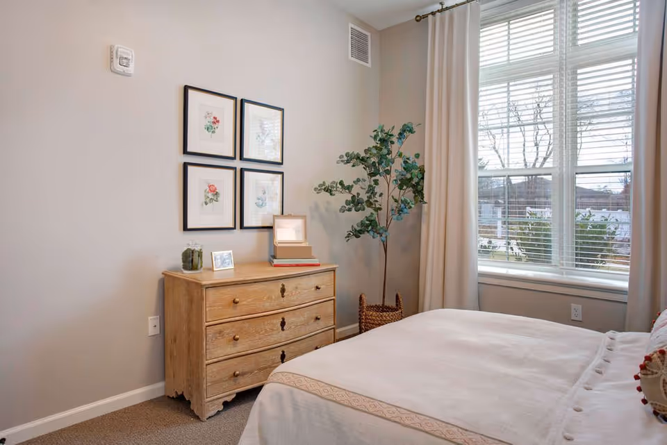 Sunlit bedroom with a bed, wooden dresser topped with decor, framed wall art, and a large window with curtains.