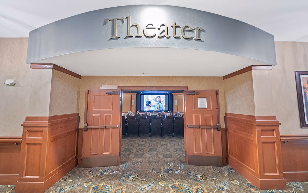 Entrance to a theater room in a senior living facility with double wooden doors open, showing rows of seated people watching a movie on a large screen. The word 'Theater' is displayed above the entrance.