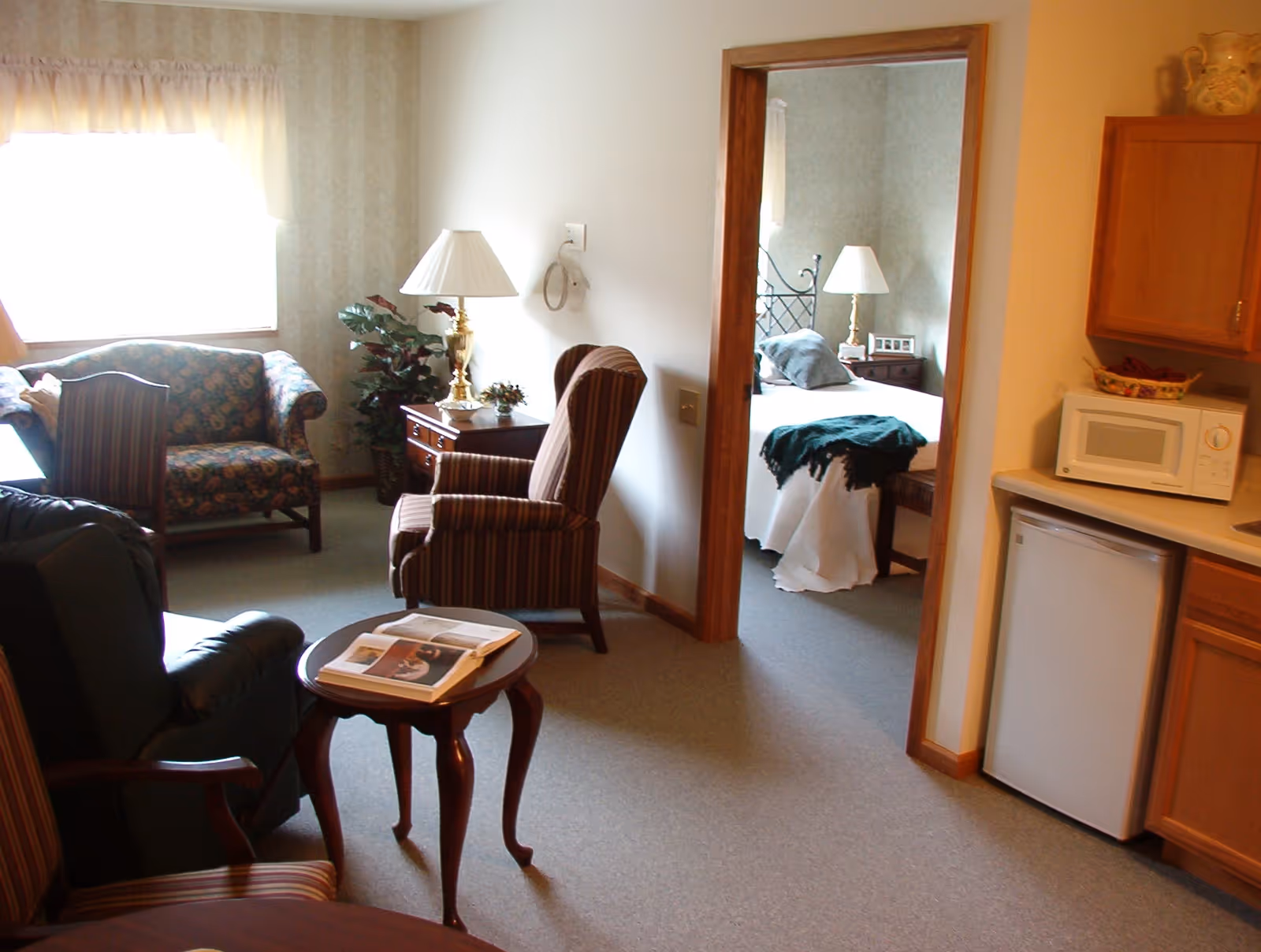 A cozy living area with a floral couch, striped armchairs, a small wooden table with an open book, and a lamp on a side table. Through an open doorway, a bedroom is visible with a bed covered in white bedding and a green throw blanket. To the right, there is a kitchenette area with a microwave, mini refrigerator, and wooden cabinets.