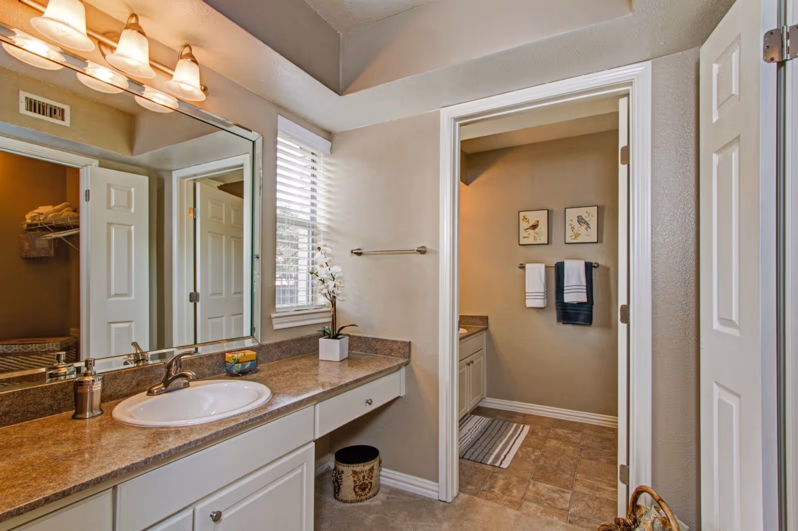 Spacious bathroom vanity with sink, large mirror and lighting, leading to an adjoining washroom with towels and wall art.
